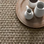 Close-up of a textured jute rug with a wooden tray and white ceramic pitchers.